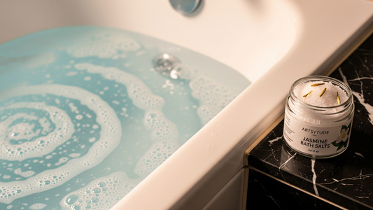 A white bathtub filled with bubbling, light blue water, alongside a jar of 'ArtsyTude Jasmine Bath Salts' on a black marble counter, suggesting a relaxing bath experience.